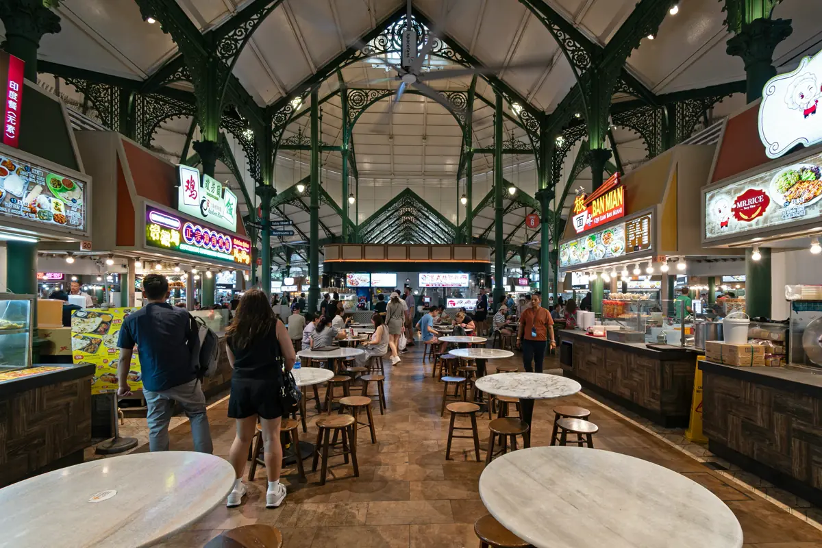 Inside a busy Singapore hawker centre, with food stalls along both sides and round tables in the middle